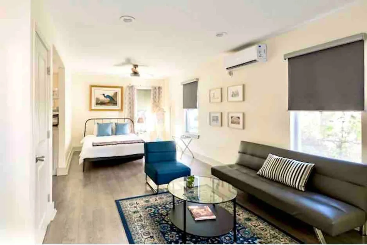 A serene, tastefully decorated living area featuring a comfy sofa, a blue armchair, a coffee table, and a plush bed in the background. Soft lighting filters through the window, setting a tranquil mood ideal for couples enjoying quiet moments or sharing stories together.