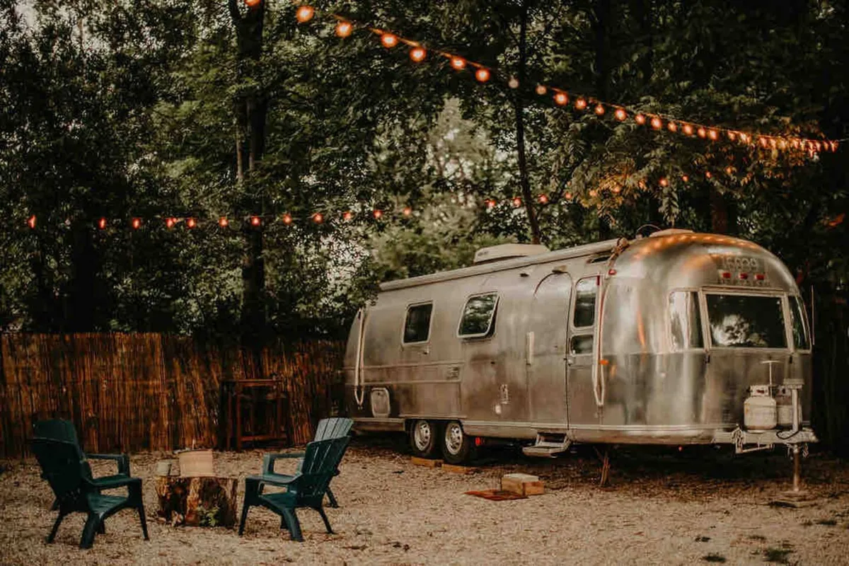 An exterior view of an Airstream trailer with a campfire, string lights, and a welcoming seating area. The scene radiates warmth and nostalgia, suggesting a unique hideaway experience connected to nature.