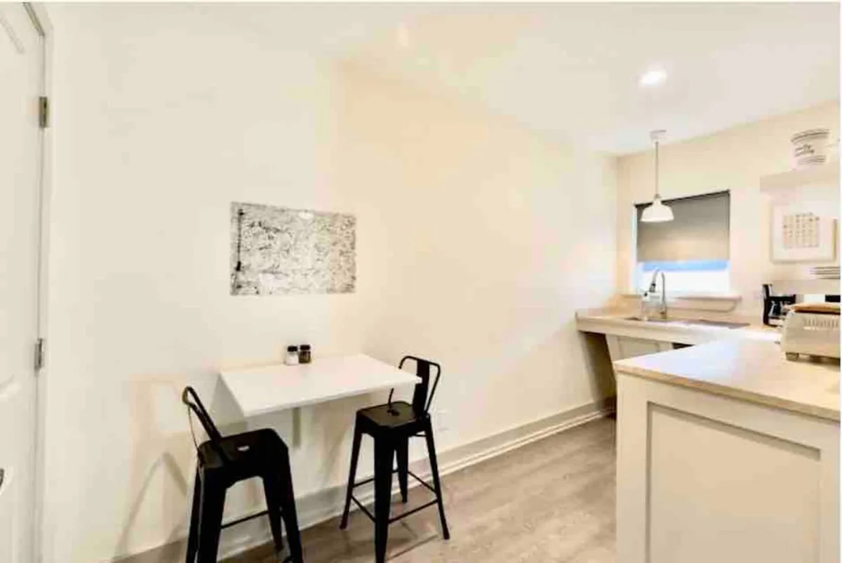 A cozy kitchenette and dining area with a small white table, two black bar stools, abstract artwork on the wall, and a kitchen countertop set for casual gatherings. The space is designed for small groups to prepare snacks, share meals, or enjoy coffee together.