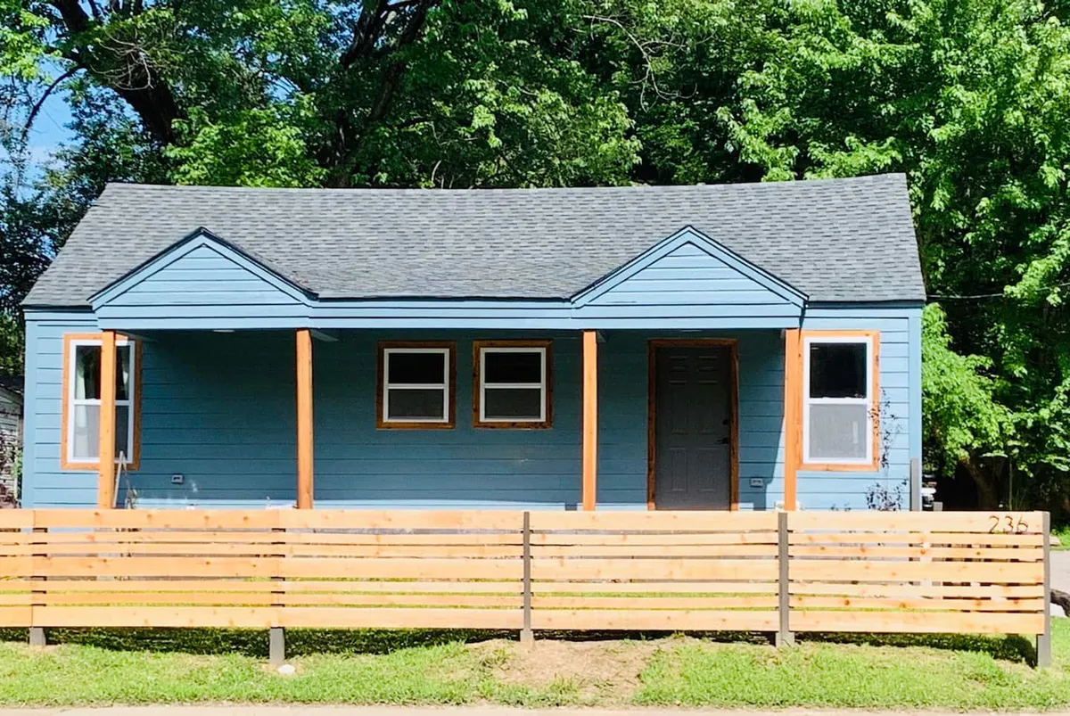 A cheerful exterior view of the blue house with a welcoming front porch, bordered by a wooden fence and surrounded by greenery. The porch invites guests to relax, and the setting hints at the neighborhood's charm and walkability.