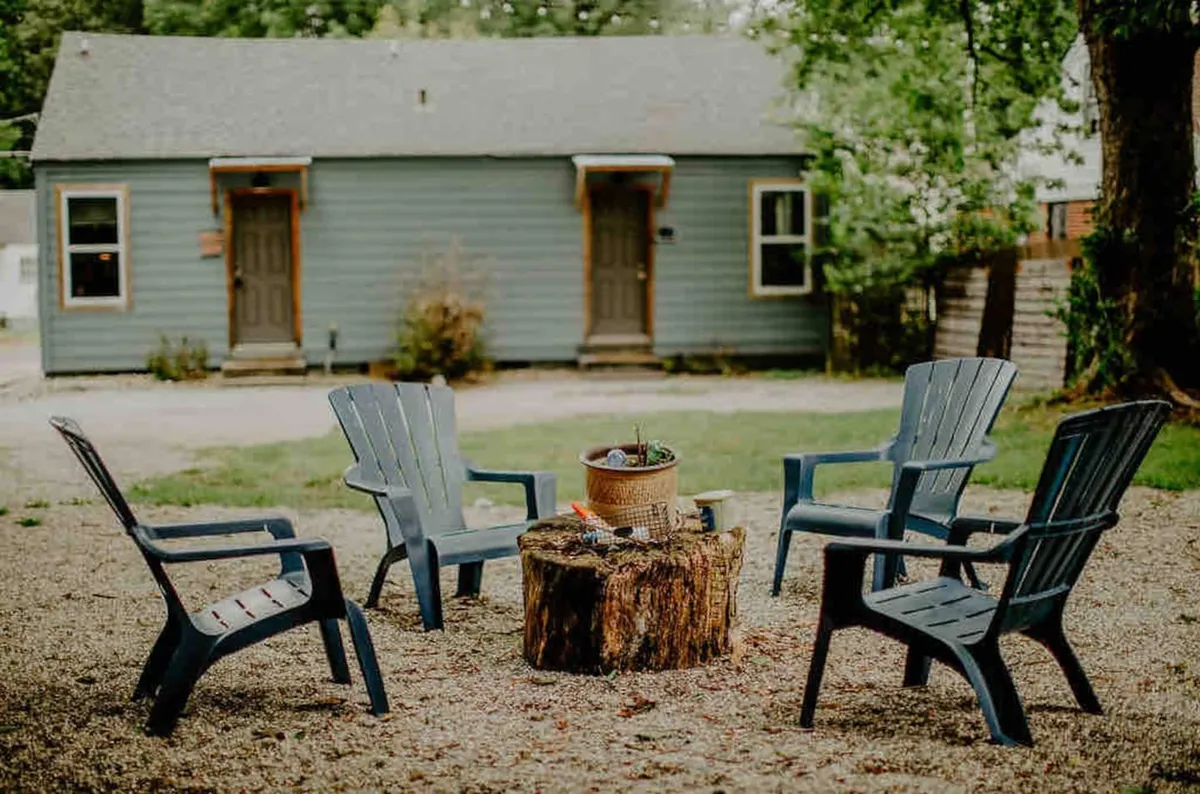 An inviting outdoor fire pit area surrounded by wooden stump seats and Adirondack chairs, set against a backdrop of greenery. This space is clearly designed for communal gatherings, offering an authentic campground vibe with the comfort of home amenities nearby.