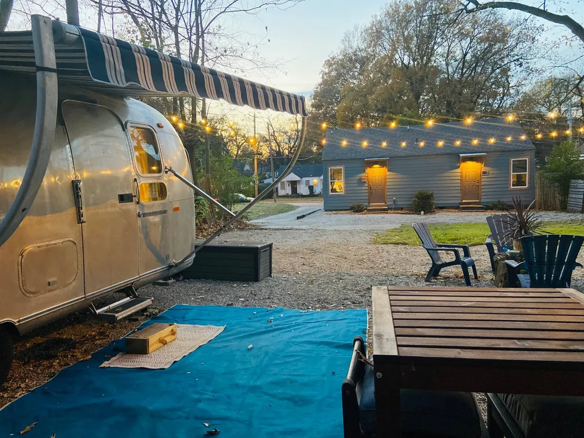 The image showcases an inviting outdoor scene featuring a gleaming Airstream trailer beneath a canopy of glowing string lights. A large wooden table surrounded by comfortable chairs suggests a welcoming space for outdoor dining and memorable gatherings. The cozy, communal setting is set against a peaceful, natural backdrop, perfect for relaxing and connecting with friends or family after a day of Memphis adventures.