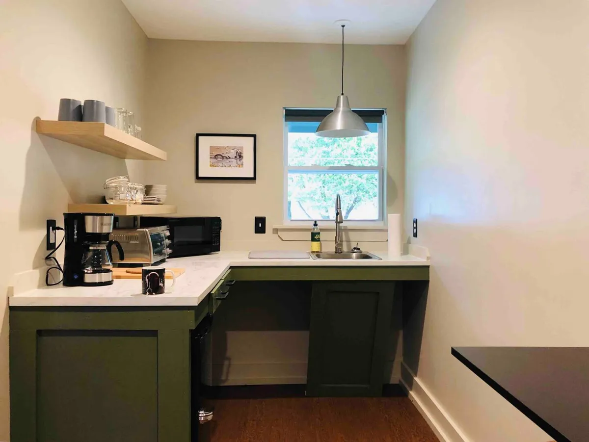 A compact and inviting kitchenette setup with a sink, coffee maker, and shelves stocked with glassware. A window brings in natural light, creating a pleasant spot for morning routines or casual meals.